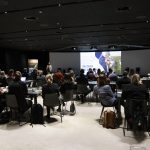 Seminaari, ihmisiä istuu ryhmissä pöytien äärellä ja kuuntelee puhujaa.